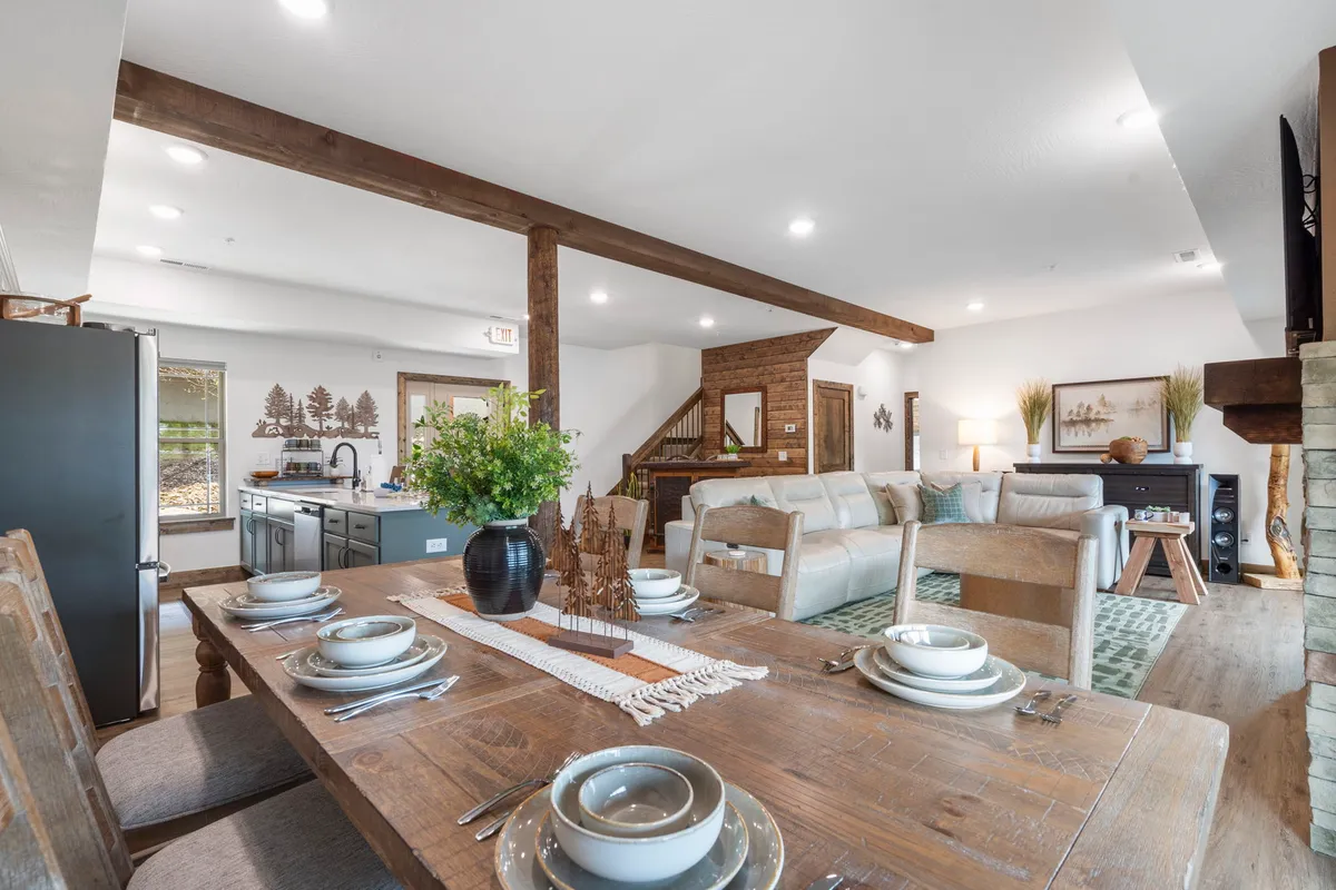 A welcoming dining area with a generously sized table set for a meal sits adjacent to a comfortable sofa. Wooden beams and decorative plants add warmth, and the scene is set for memorable group feasts or relaxed evenings.