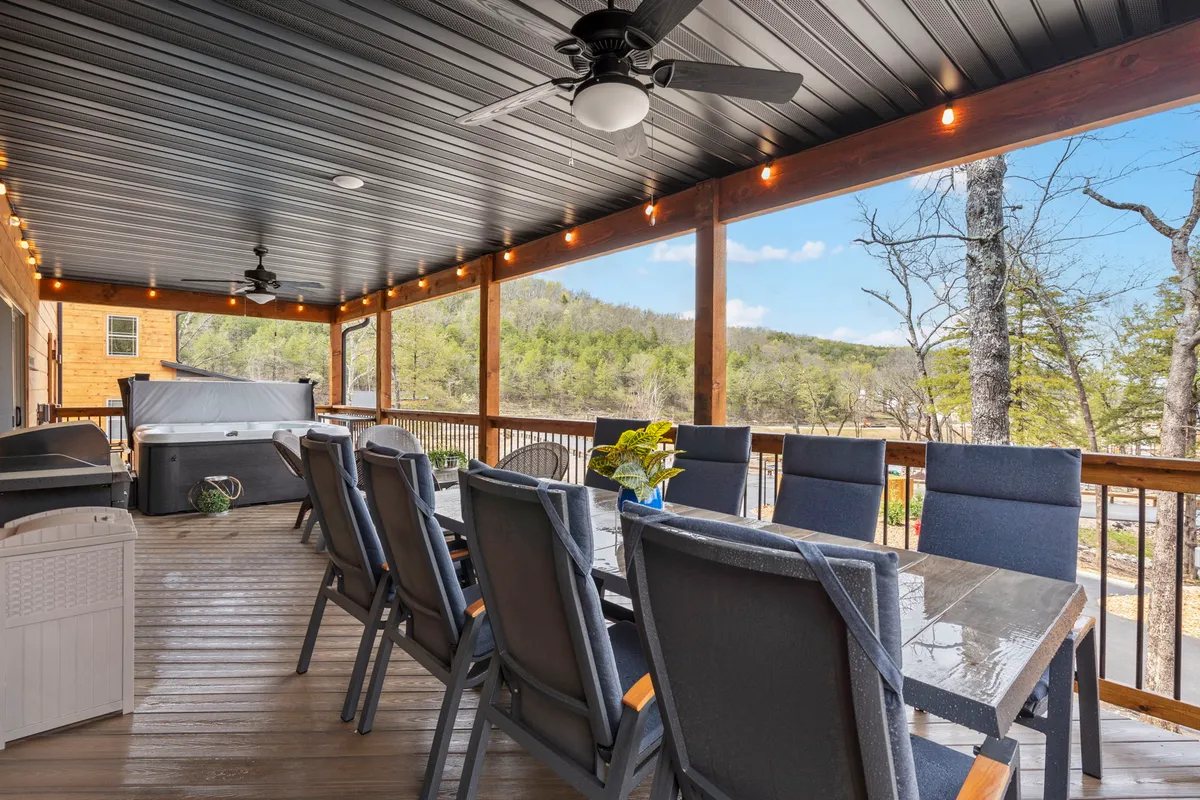 A spacious outdoor area featuring a large dining table surrounded by comfortable seating, with a bubbling hot tub nearby. The backdrop of rolling hills and lush greenery creates an inviting environment for gatherings. The natural landscape view enhances the sense of retreat and relaxation.