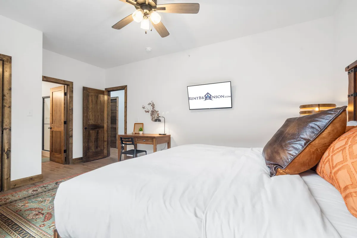 A serene bedroom featuring a plush bed adorned with decorative pillows, a cozy desk perfect for reading or working, a mounted television for entertainment, and elegant wooden doors that add warmth to the space.