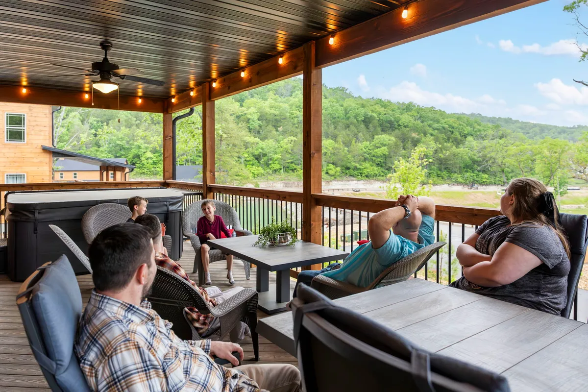 An inviting outdoor seating area featuring a hot tub, surrounded by lush greenery and a spacious deck. People gather around a table enjoying the outdoors, with the landscape offering a tranquil backdrop for both relaxation and socializing after a day of exploring.