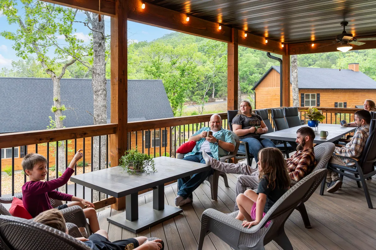 A wooden deck set against a mountain backdrop, illuminated by string lights overhead. Groups of people are sitting and interacting at tables and lounge chairs, with games and celebratory meals taking place. The atmosphere is lively yet relaxed, perfect for gatherings at sunset.