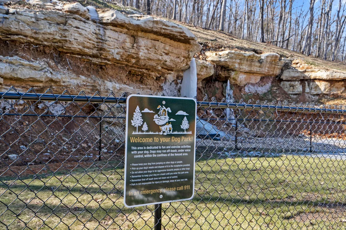 A cheerful dog park with a prominent sign, rock formations, and a securely fenced area. The space is clearly designed for dogs to play safely, and the natural surroundings create a welcoming environment for pet owners and their furry companions.
