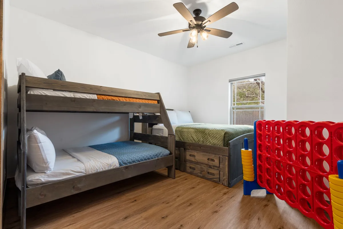 A playful bunk room showcasing a twin-over-full bunk bed with a vibrant green bedspread, a queen-sized bed, and a red Connect Four game, creating a fun and inviting space for kids or groups of friends.