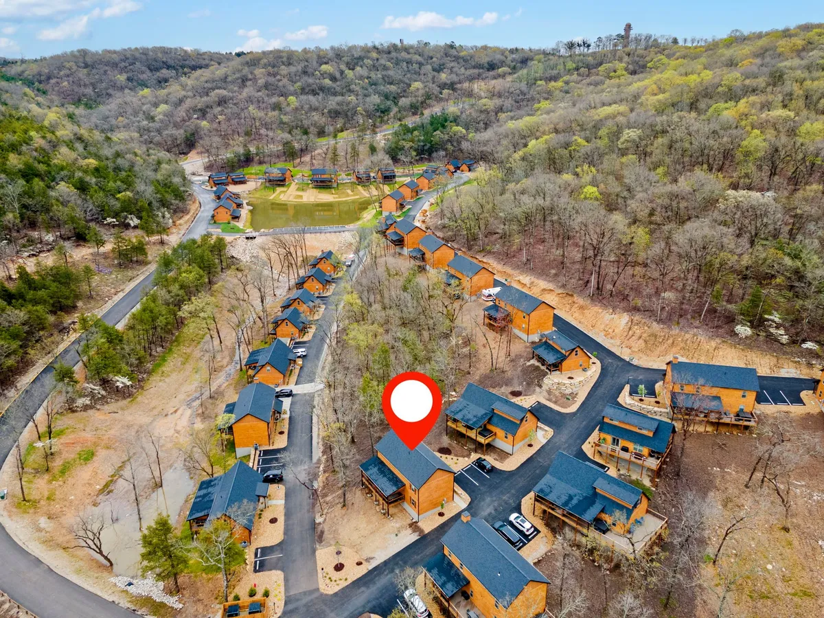 A serene cluster of cozy cabins nestled among trees, with a central pond reflecting the greenery and sky. Gentle trails wind between the cabins, inviting exploration and nature walks. The presence of the pond hints at opportunities for quiet reflection, fishing, or picnics by the water.