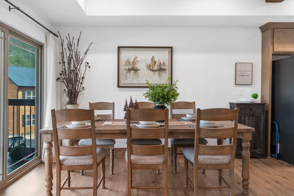 The image showcases a spacious interior dining area centered around a sturdy wooden dining table, adorned with a stylish greenery centerpiece and decorative branches in a vase. Framed artwork adds character to the walls, while natural light fills the room, creating a warm, inviting atmosphere ideal for group gatherings and family meals.
