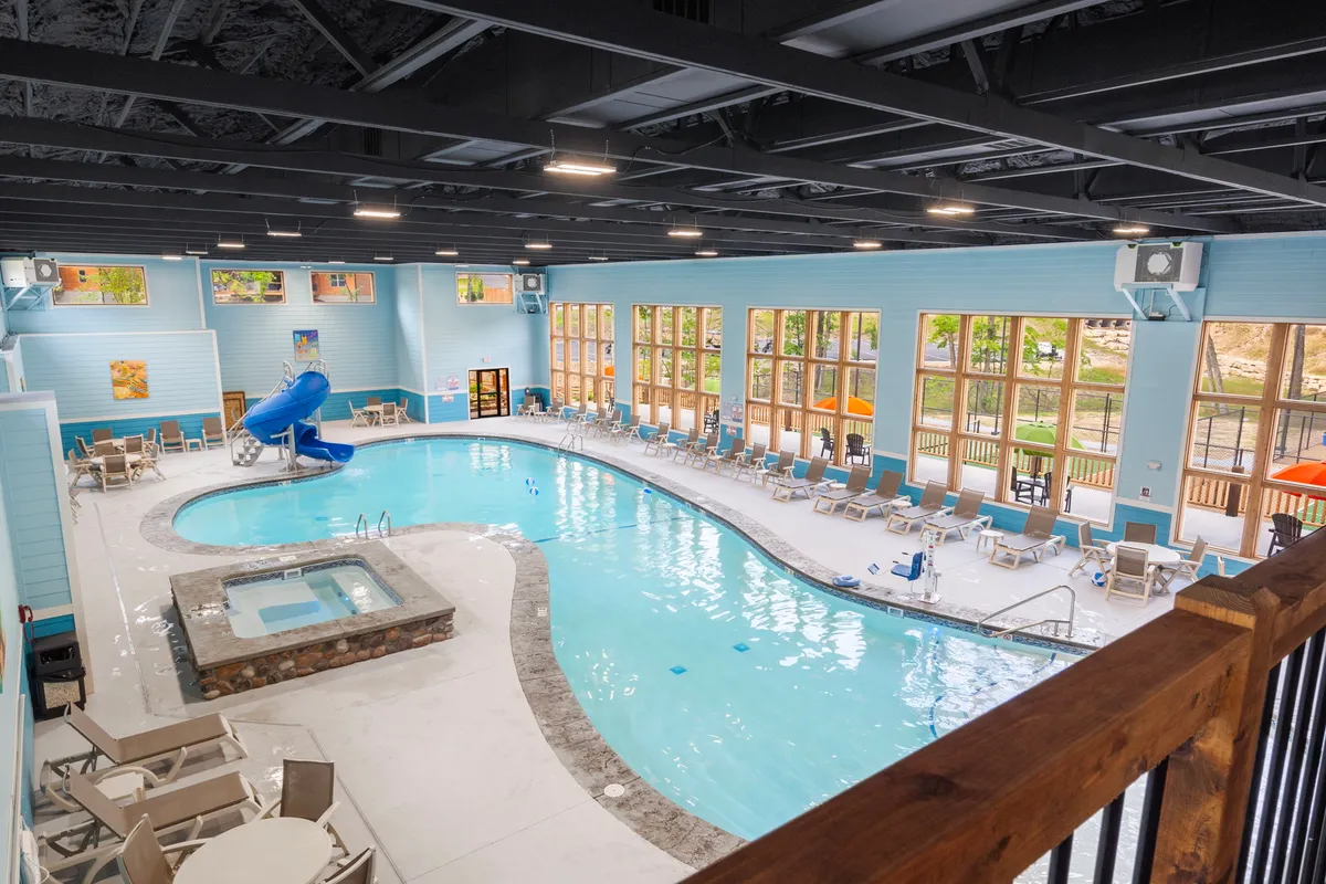 A vibrant indoor pool area featuring a water slide, hot tub, lounge chairs, and large windows letting in sunlight. People are enjoying the relaxing and playful atmosphere, highlighting the resort’s year-round aquatic amenities.